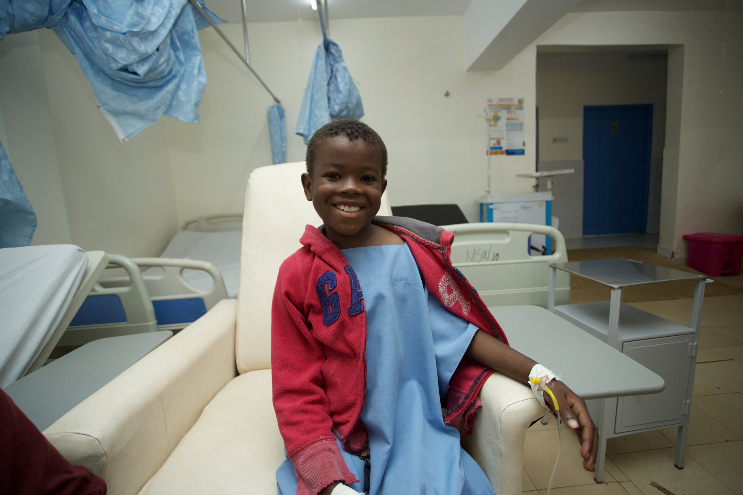 Dennis sits smiling in a treatment chair at Chogoria Hospital, an IV line taped to his hand.