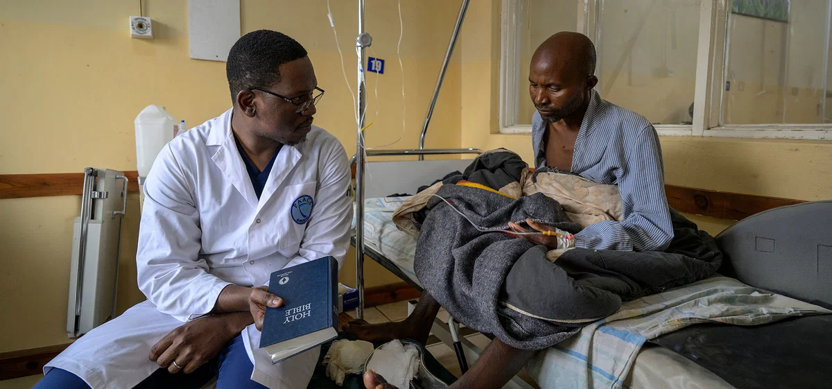 A doctor in a white coat holds a Bible as he sits and talks with a patient beside his hospital bed.