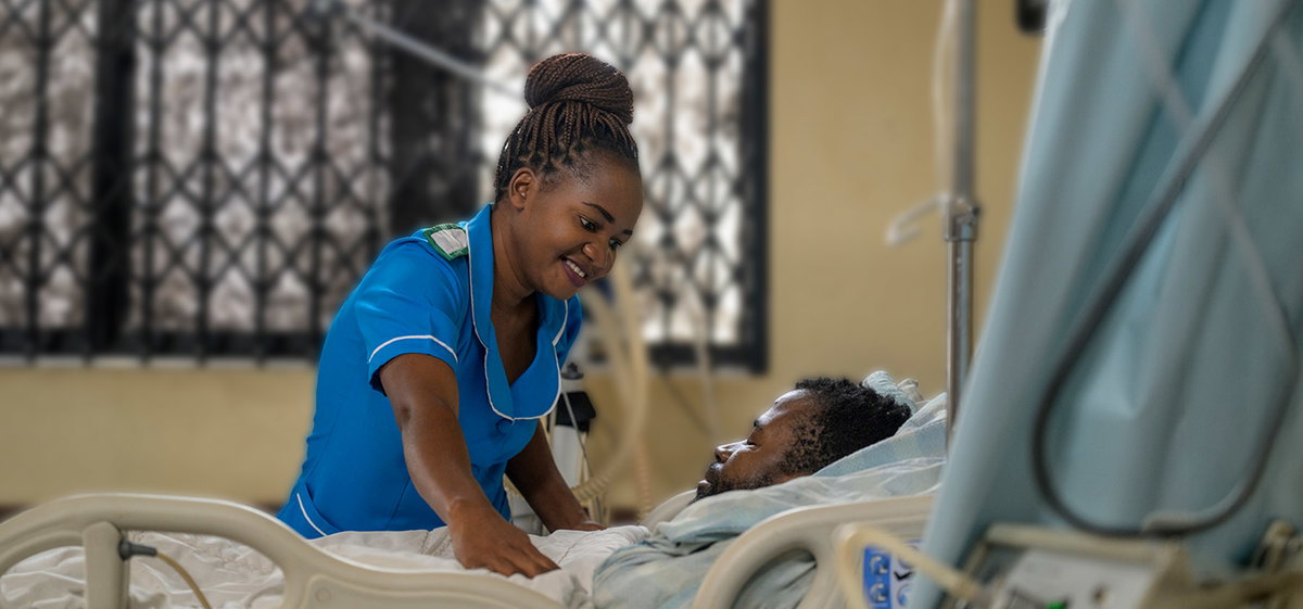 A smiling nurse in blue scrubs leans over a patient resting in a hospital bed.