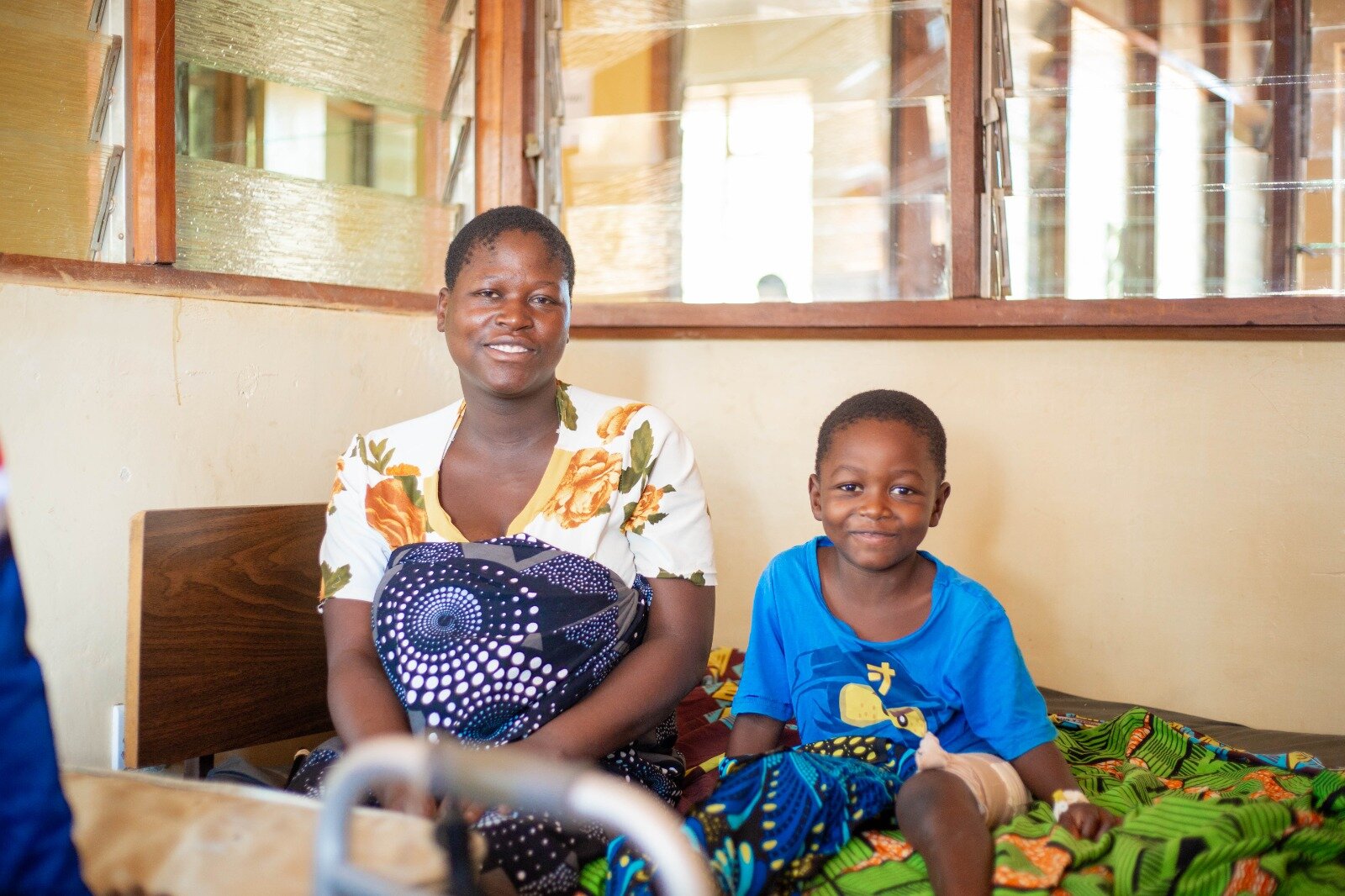 Reuben smiles from a hospital bed at Ekwendeni Mission Hospital, his leg bandaged, beside his mother.